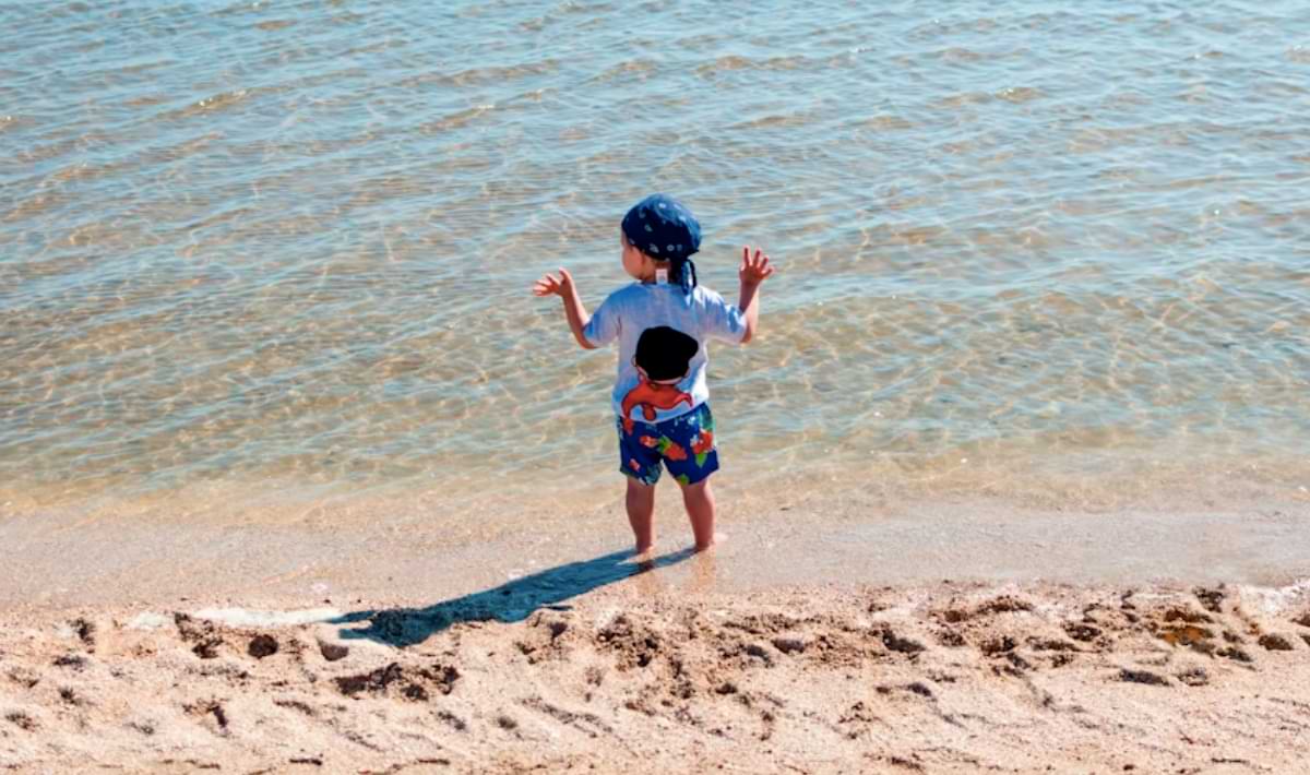 Small child in a blue hat and colorful swim trunks stands at the edge of clear, shallow water on a sandy beach, watching gentle waves lap at their feet.