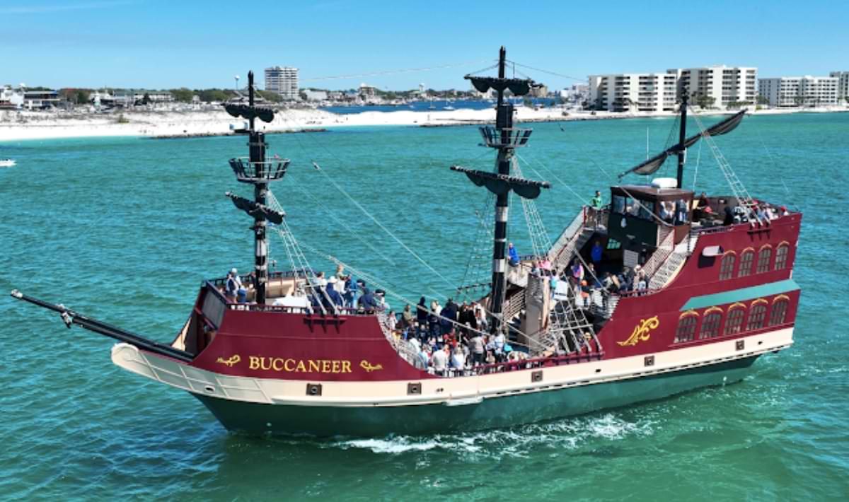 A large red and cream pirate-themed tour boat labeled “Buccaneer” sails through bright emerald water, with passengers on deck and Destin’s white-sand shoreline and condos in the background.