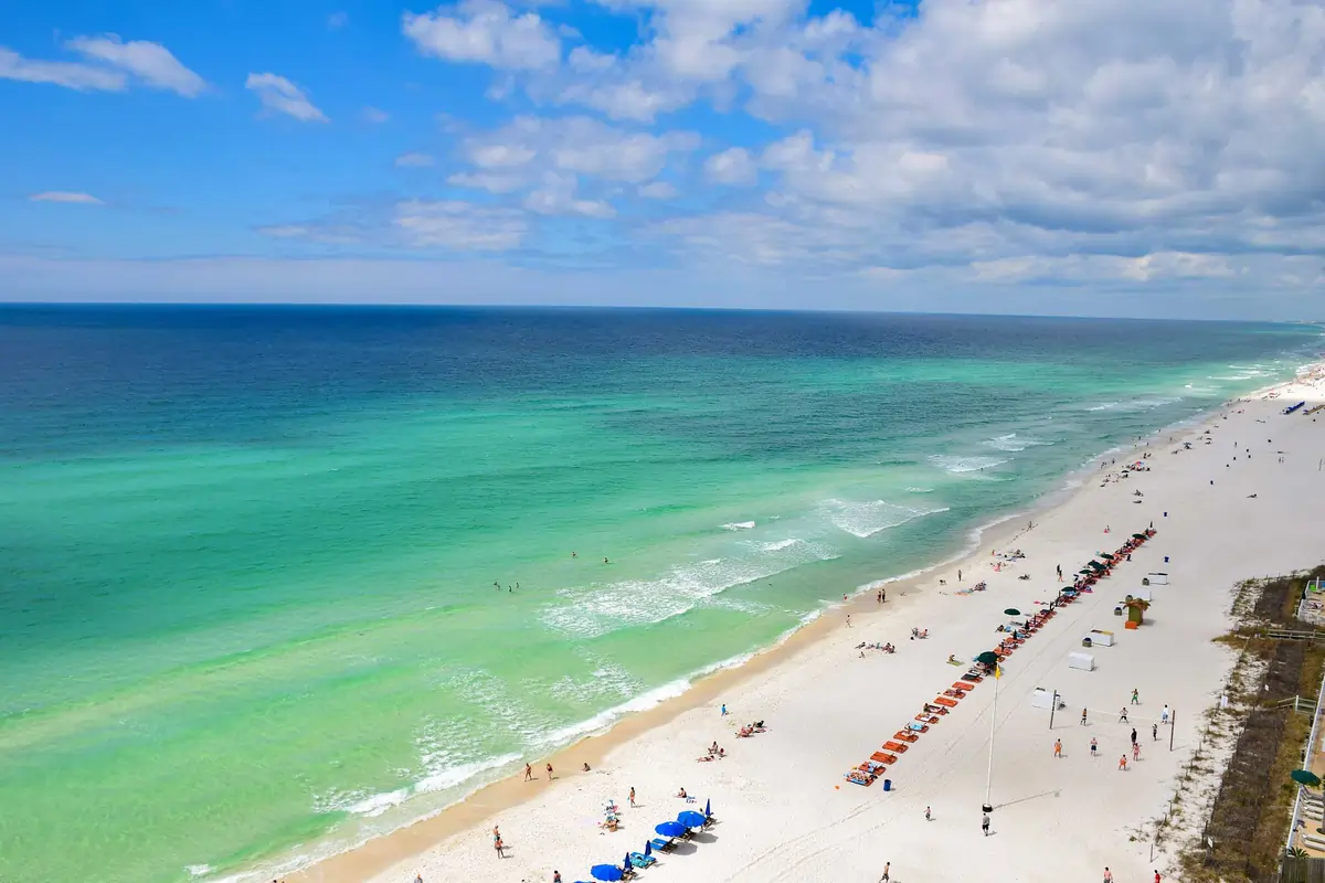Destin, Florida shoreline with calm emerald Gulf water, sugar-white sand, rows of beachfront condos, and neatly arranged beach chairs along the water’s edge.