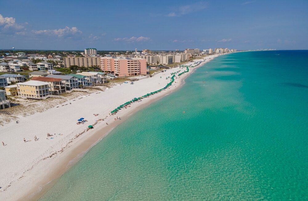 Aerial view of Destin, Florida with emerald-green Gulf water, sugar-white sand, rows of beachfront condos, and a line of green beach umbrellas along the shoreline.