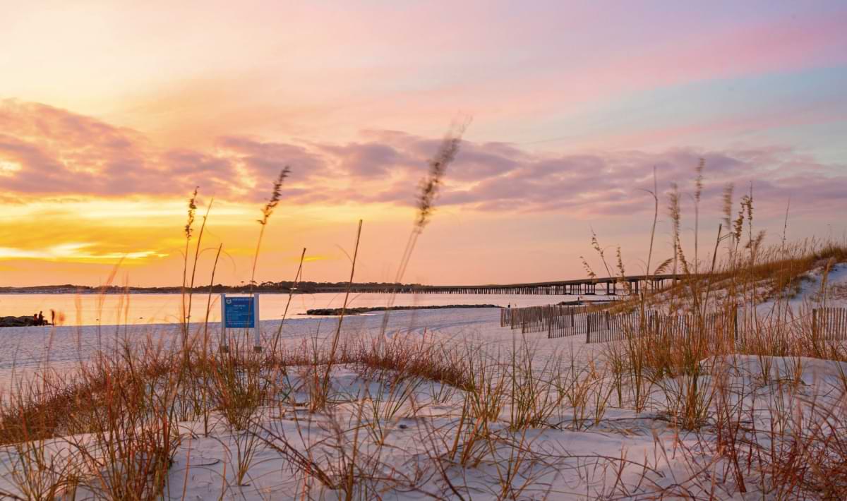 Soft sunset light glows over white sand dunes and sea oats, with calm water, a distant pier, and pastel clouds filling the sky at a quiet Destin, Florida beach.