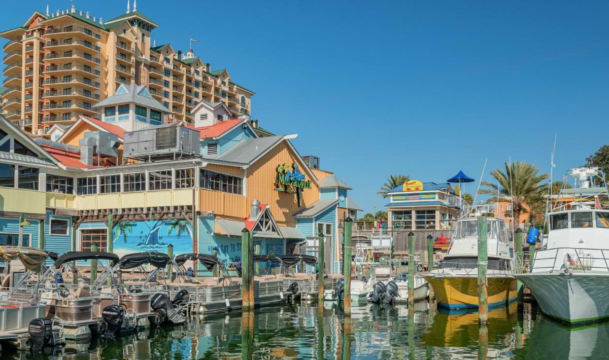 Colorful waterfront buildings and restaurants at Destin Harbor overlook rows of docked boats and charter vessels on a sunny day with a clear blue sky.