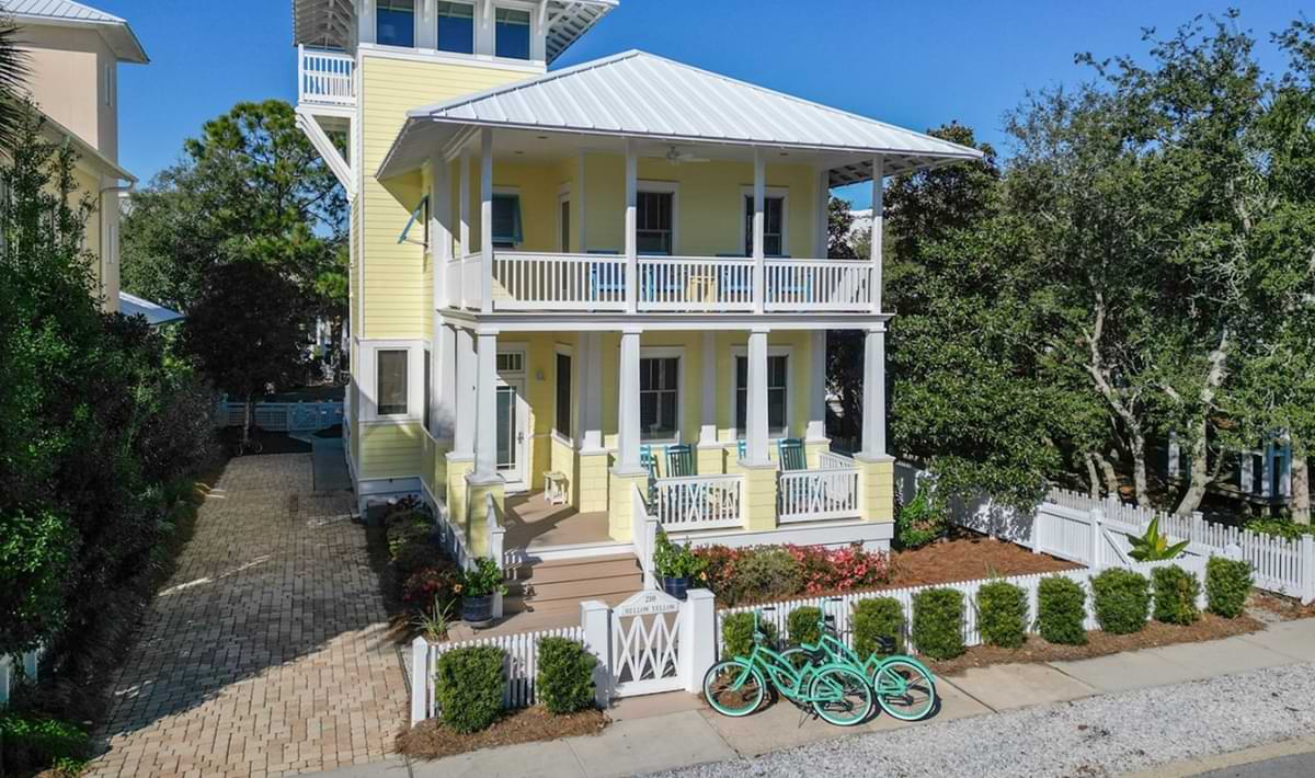 Two-story yellow beach cottage with wraparound porches, white trim, and a picket fence, featuring a brick driveway and several turquoise cruiser bikes lined up along the front sidewalk.
