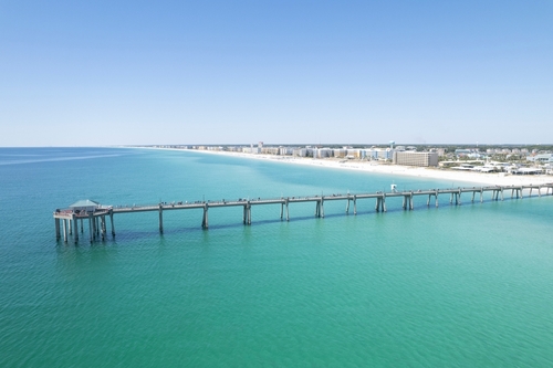 Aerial view of the Fort Walton Beach fishing pier stretching over calm emerald-green Gulf water with white-sand shoreline and beachfront resorts in the distance.