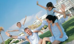 Two adults sit on a sunny resort lawn with two children balancing playfully on their shoulders, all wearing light blue outfits and stretching their arms out as they laugh and pose together.