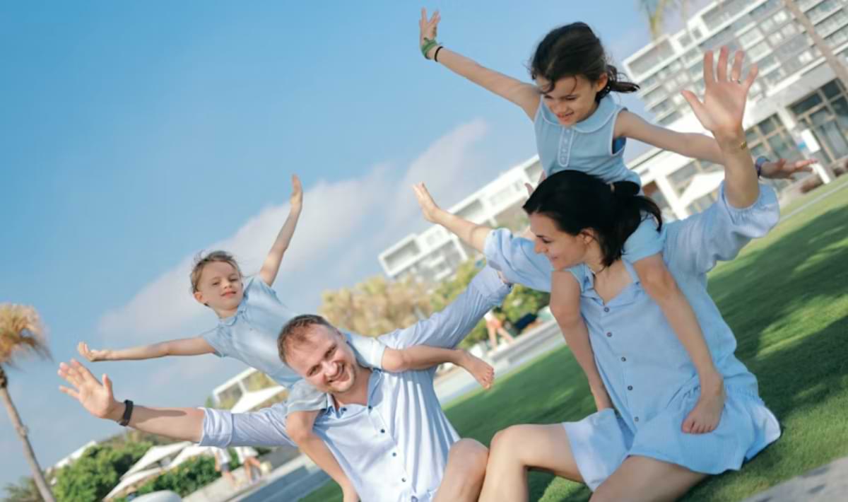 Two adults sit on a sunny resort lawn with two children balancing playfully on their shoulders, all wearing light blue outfits and stretching their arms out as they laugh and pose together.