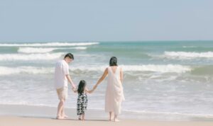 Two adults and a child holding hands walk barefoot at the edge of the surf on a sunny beach, facing gentle waves rolling in from the turquoise water.