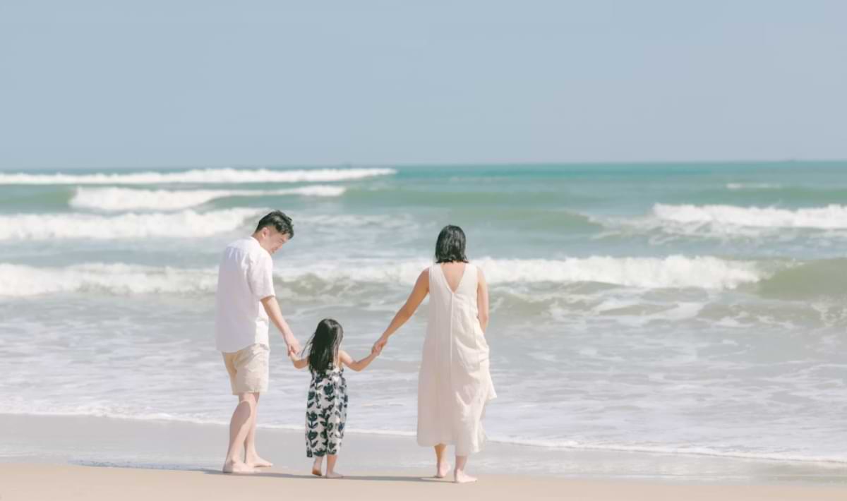 Two adults and a child holding hands walk barefoot at the edge of the surf on a sunny beach, facing gentle waves rolling in from the turquoise water.