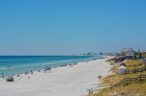 Miramar Beach shoreline with calm emerald water, soft white sand, scattered beachgoers, and a mix of gulf-front homes and distant high-rise condos along the coast.