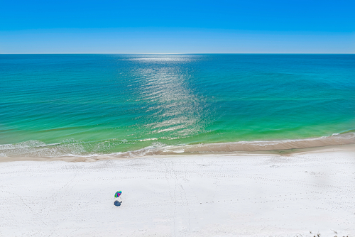 Panama City Beach shoreline with calm emerald-green Gulf water, sugar-white sand, and a single colorful beach umbrella on an uncrowded stretch of coast.