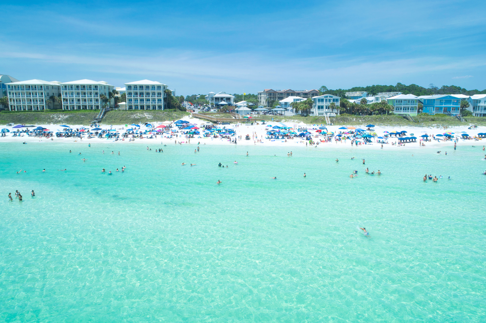 Santa Rosa Beach aerial with clear turquoise Gulf water, white sand shoreline, and beachfront condos and homes behind rows of colorful umbrellas and swimmers.