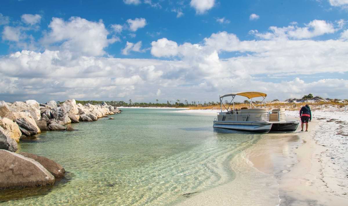 A pontoon boat with a yellow canopy is pulled onto the clear shallow water of Shell Island, beside white sand shoreline and a rocky jetty under a bright blue sky with scattered clouds.