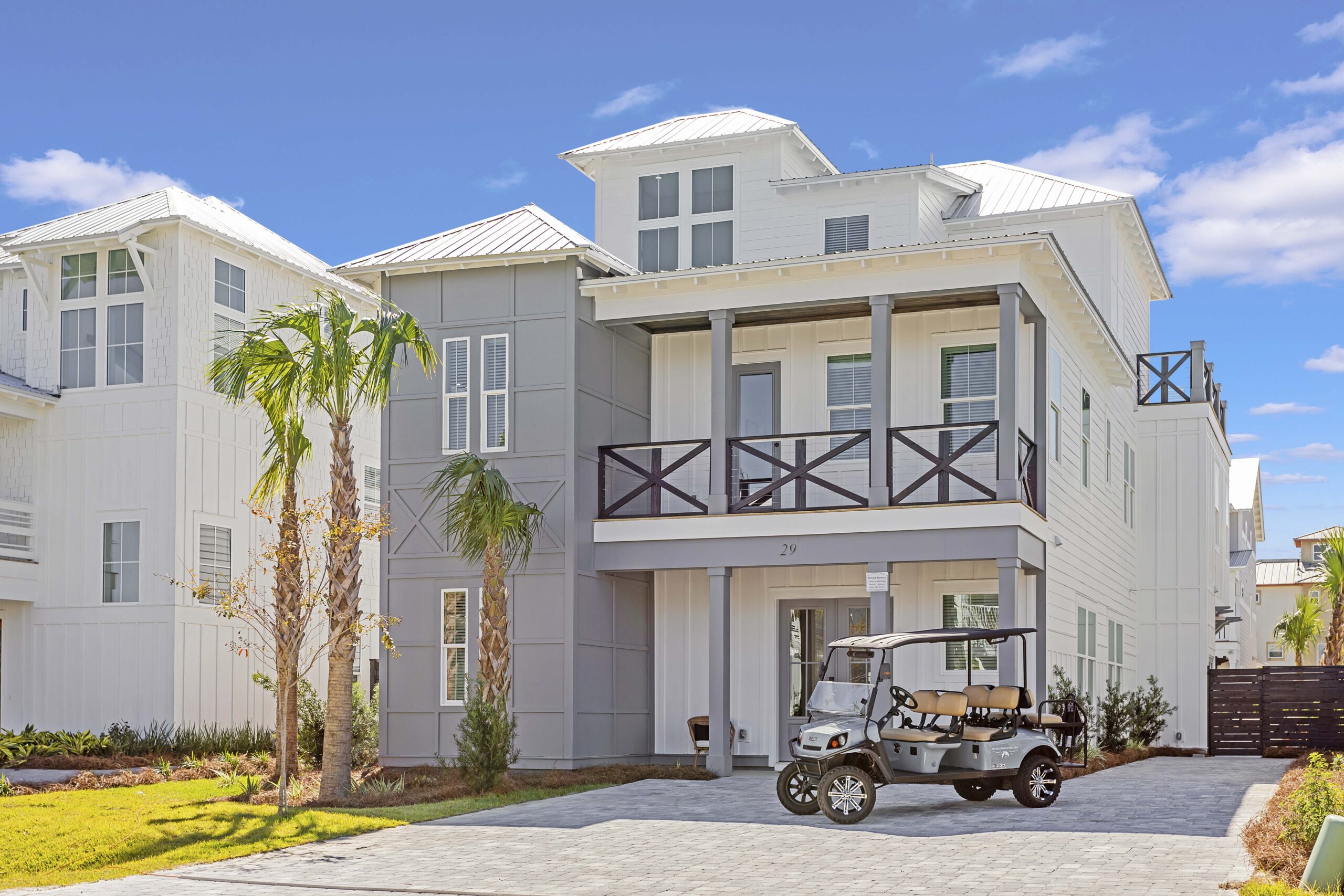 Front view of a modern three-story coastal home with white siding, gray accents, and a second-story balcony, featuring palm trees in the yard and a parked golf cart in the driveway on a sunny day.