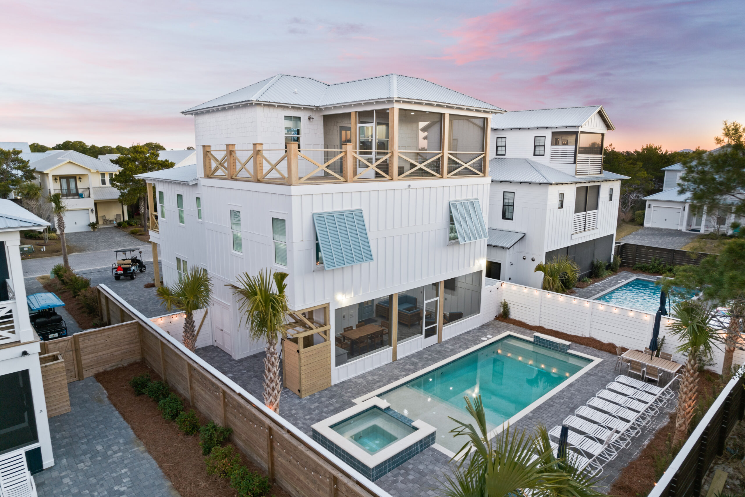 A modern three-story white beach house with blue shutters features a fenced backyard with a rectangular pool, spa, lounge chairs, and palm trees, set beneath a colorful sunset sky in a coastal neighborhood.