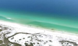 Aerial view of Topsail Hill Preserve State Park with bright white sand dunes, scattered greenery, and calm turquoise Gulf waters fading into deep blue offshore.