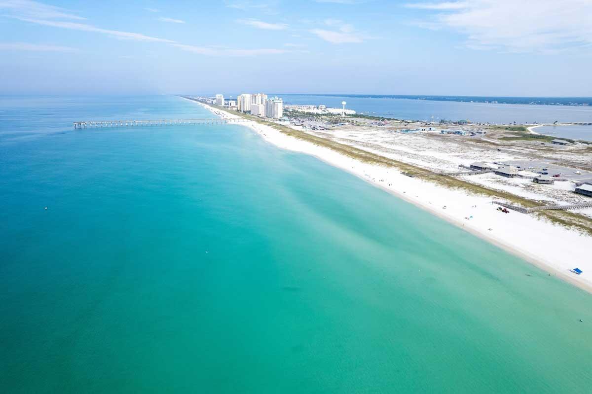 Navarre Beach aerial with calm turquoise Gulf water, long white-sand shoreline, fishing pier, and a row of beachfront condos and dunes along the coast.